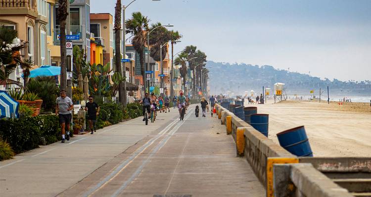 Drukke boulevard aan het strand met fietsers, joggers en kleurrijke huizen naast een zandige kust.