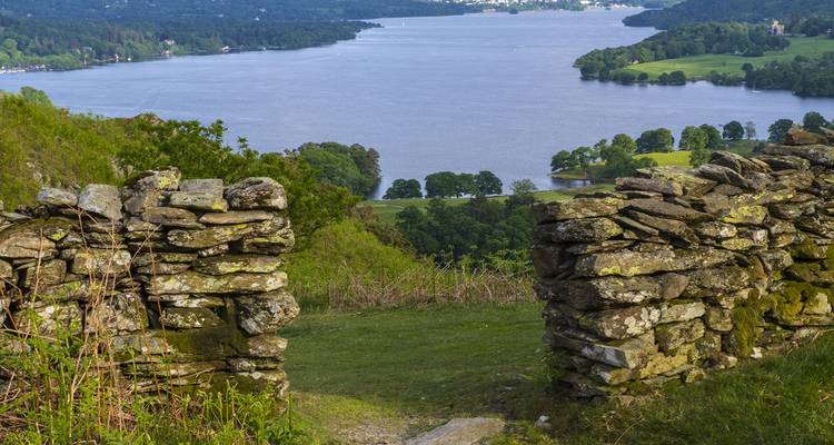 Sereen uitzicht over Loch Lomond omlijst door stenen muren en glooiende groene schiereilanden onder helderblauwe hemel.