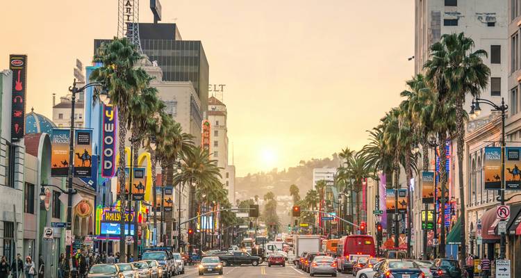 Resplandor del atardecer sobre un Hollywood Boulevard concurrido bordeado de palmeras, tráfico y vallas publicitarias.