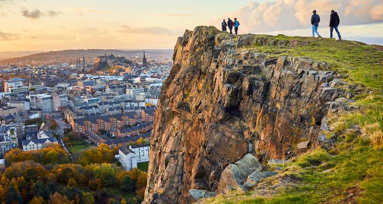 Wandelaars die bovenop een rotsachtige klif staan en uitkijken over de skyline van Edinburgh tijdens het gouden uur.