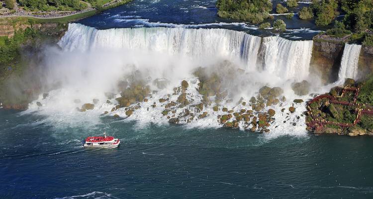 Vista aérea de las Cataratas del Niágara con neblina elevándose, base rocosa visible y barco turístico acercándose a la cascada.