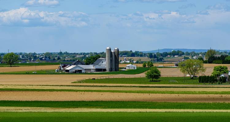 Paisaje rural pacífico del Condado de Lancaster con campos de cultivo verdes y marrones, graneros y silos bajo un cielo parcialmente nublado.