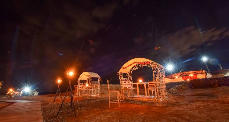 Escena nocturna de campamento en el desierto con carromatos blancos ornamentados y llamas de antorchas bajo un cielo estrellado.