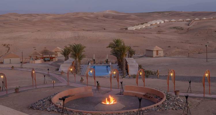 Panorama amplio de campamento en el desierto con fogata central, palmeras y pequeña piscina al atardecer.