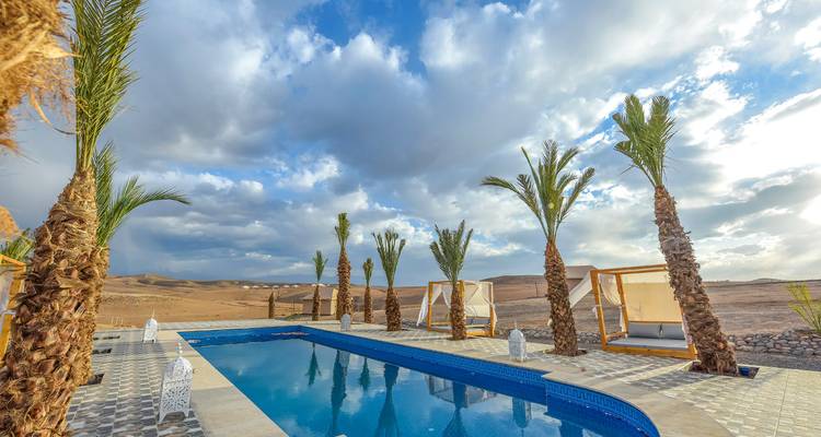 Piscina al aire libre rodeada de palmeras jóvenes con vista a extensos paisajes desérticos bajo un cielo dramático.