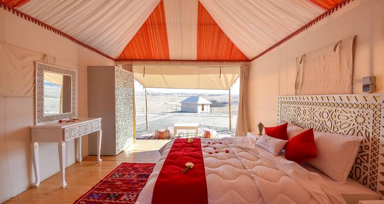Interior acogedor de tienda con acentos rojos, cama bien hecha y vista del desierto a través de las paredes abiertas de lona.