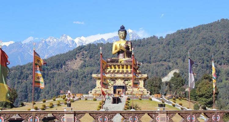 Grande statue dorée de Bouddha assise sur une plateforme ornée avec des sommets enneigés en arrière-plan.