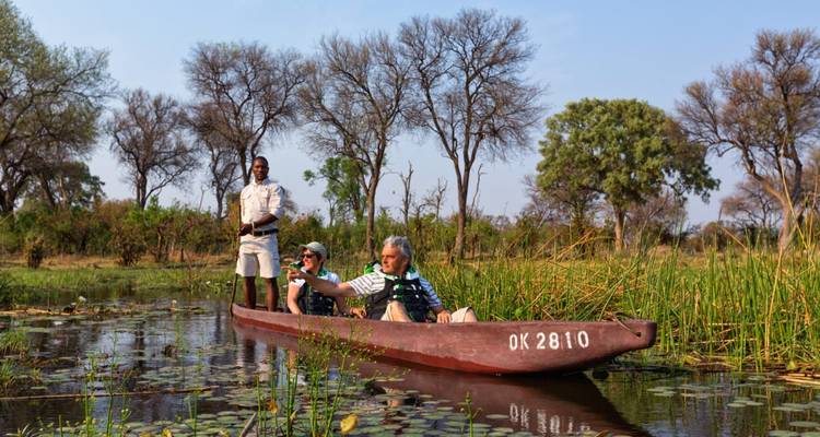 Guide standing on a mokoro while two tourists sit photographing wildlife in a calm delta channel lined with reeds and lilies.