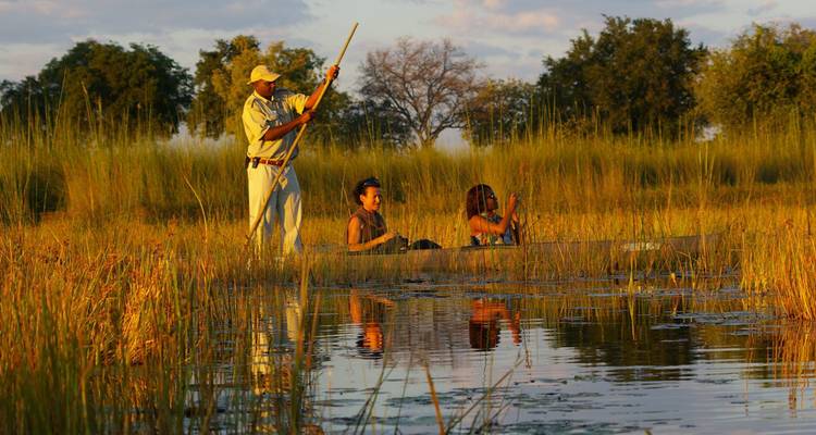 Three travelers paddling a wooden canoe through tall reeds in warm evening light on the Okavango.