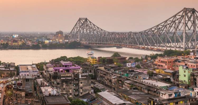 Howrah Bridge spans the Hooghly River at dusk above the colorful rooftops of Kolkata.