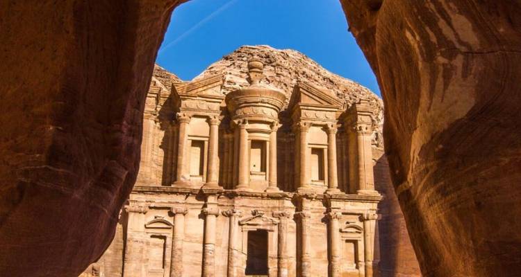 Vue du Monastère de Pétra (Ad-Deir) à travers une arche naturelle de grès sur fond de ciel bleu profond.
