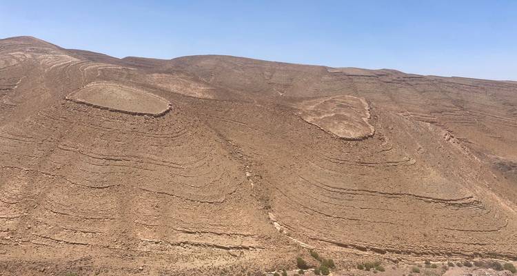 Schichtige, trockene Gesteinsformationen, die schwungvolle Muster an einem kargen Berghang unter blassblauem Himmel bilden