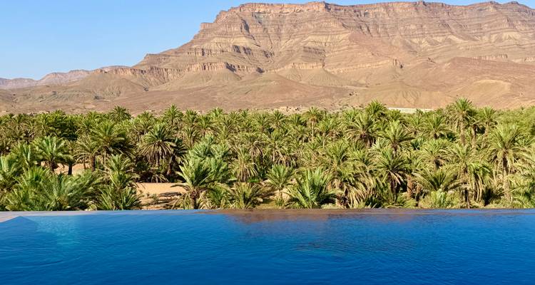 Une piscine à débordement surplombe une dense oasis de palmiers encadrée par des montagnes désertiques escarpées sous un ciel bleu clair.