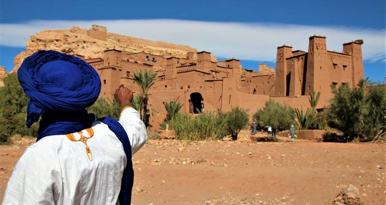 Homme au turban bleu pointe vers la kasbah en adobe d'Aït-ben-Haddou sous un ciel lumineux