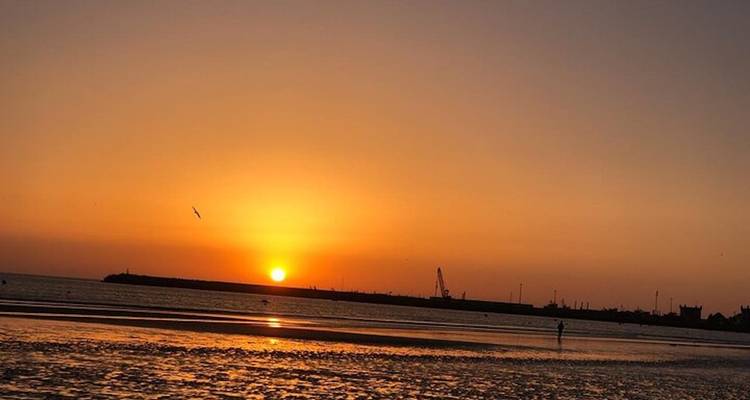 Un brillante sol anaranjado se pone sobre una tranquila costa marroquí y un puerto distante.
