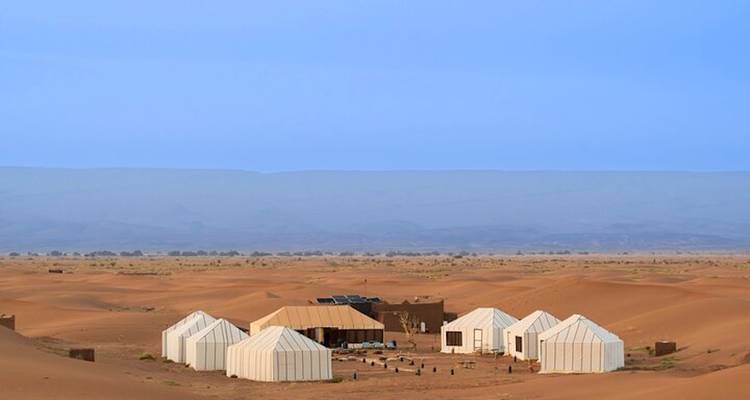 Campamento de lujo aislado con tiendas de campaña ubicado entre enormes dunas de arena naranja del Sahara bajo un cielo azul despejado.