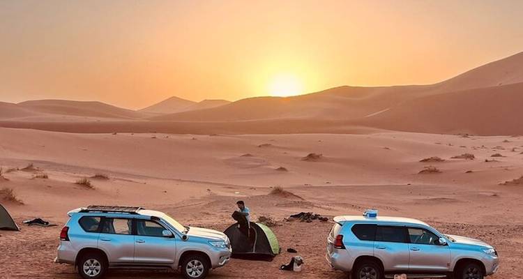 Dos vehículos 4x4 estacionados al lado de una carpa pequeña al amanecer con dunas de arena brillante de fondo.