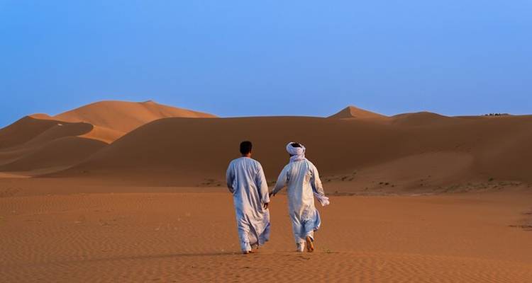 Dos hombres con ropas tradicionales caminan a través de las arenas desiertas abiertas hacia dunas altas.