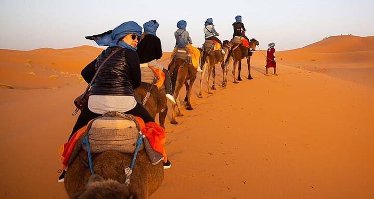 Los turistas que usan turbantes azules montan una caravana de camellos cruzando la arena dorada del desierto al amanecer.