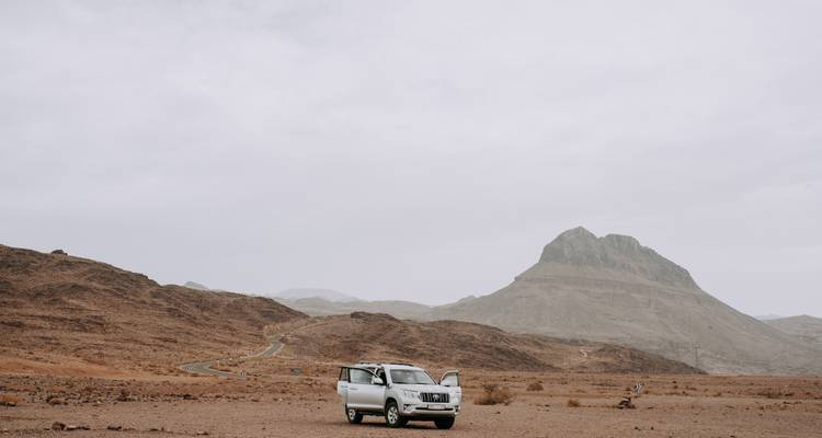 Un vehículo 4x4 solitario estacionado en una llanura desértica árida con una pico montañoso escarpado al fondo.