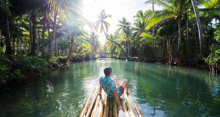 Reiziger leunt achterover op bamboevlot die door weelderige met palmen omzoomde rivierkloof drijft.