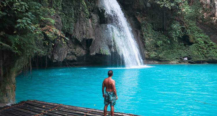 Man staat op bamboevlot gericht naar levendig turquoise watervalpoel in junglekloof.