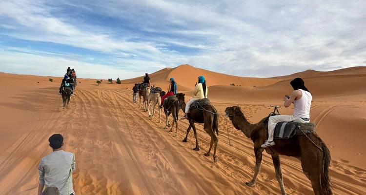 Caravana de turistas monta camellos a lo largo de dunas naranjas onduladas bajo un cielo desértico parcialmente nublado.