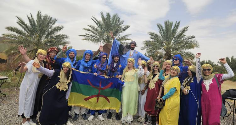 Gran grupo vestido con coloridos trajes bereberes posa alegremente con una bandera amazigh entre palmeras.