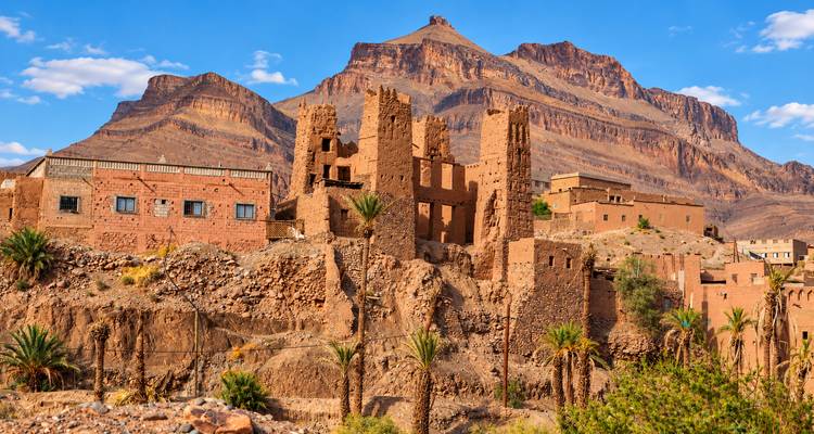 Torres de tierra arruinadas encaramadas en una ladera rocosa con un espectacular telón de fondo de montañas y palmeras dispersas.