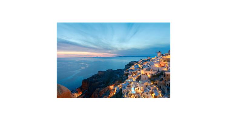 Vista nocturna de las luces del pueblo en el acantilado de Santorini contra un cielo azul tranquilo del atardecer, dentro de un marco blanco.