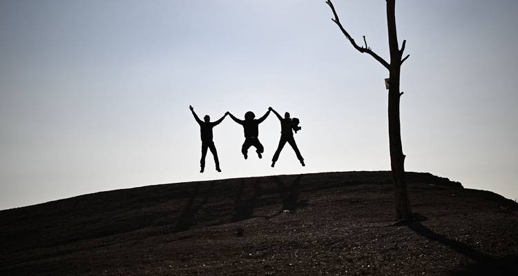 Drei Reisende, die sich an den Händen halten und als Silhouetten auf einem dunklen Hügel mit einem einsamen Ast springen.