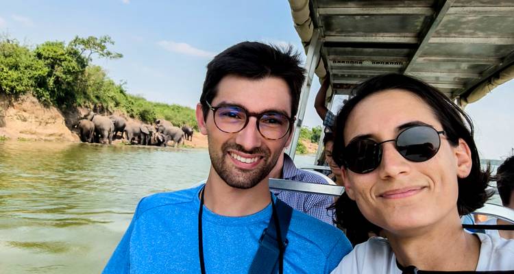 Pareja tomándose una selfie en un barco mientras elefantes se congregan en la orilla del río detrás de ellos