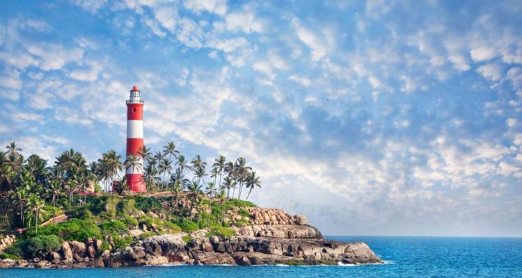 Ein rot-weißer Leuchtturm erhebt sich über Palmen auf einem felsigen Kap vor dramatischem Himmel und blauem Meer.