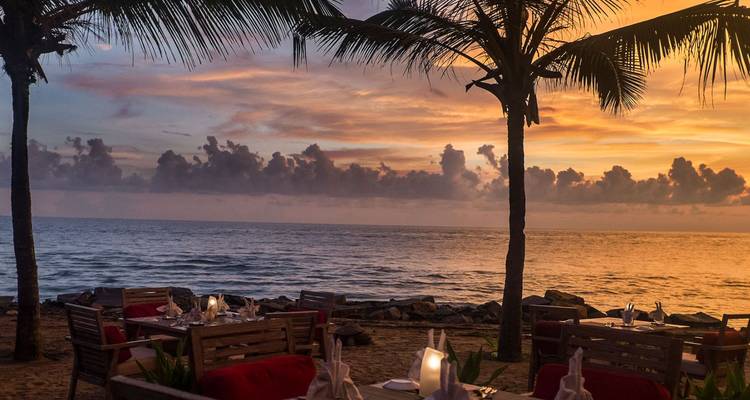Strandrestaurant-Tische unter Palmenschatten blicken auf einen farbenfrohen Sonnenuntergangshimmel.