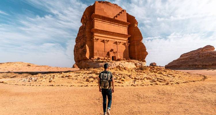 A lone traveler stands before a monumental red-sandstone tomb in a desert landscape.