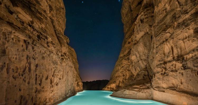 A glowing turquoise pool snakes between towering canyon walls beneath a starry night sky.