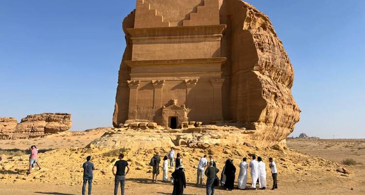 A tour group gathers in front of a monumental desert tomb carved from sandstone.