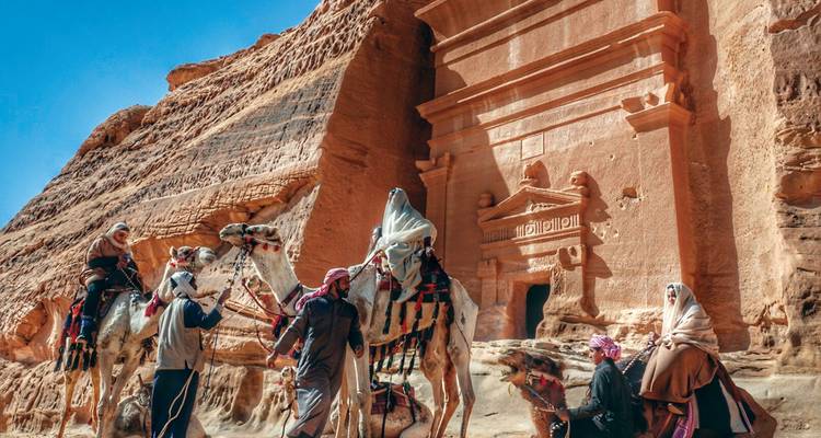 Riders on camels gather beside an ornate desert tomb carved into red rock.