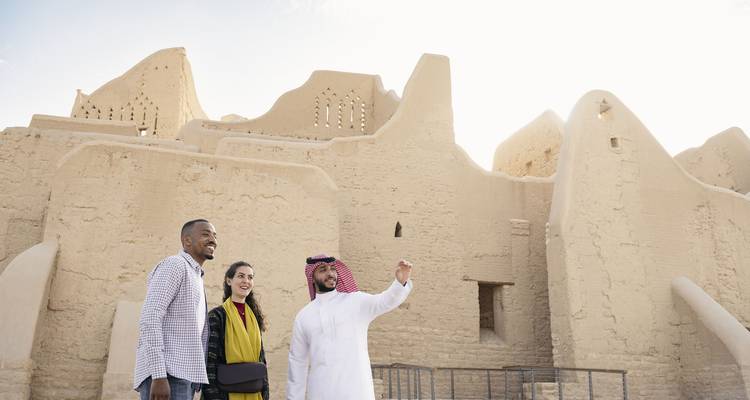 A guide shows two visitors earthen heritage buildings bathed in bright sun.