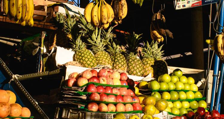 Stacks of colorful fruit—bananas, pineapples, apples, and limes—fill a sunlit market stall.