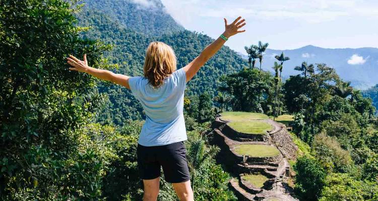Un randonneur lève les bras triomphalement en surplombant d'anciennes terrasses de pierre dans des collines de jungle dense.