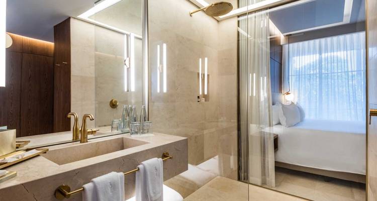Une salle de bain d'hôtel moderne avec des finitions en marbre, des accessoires dorés, et un aperçu d'une chambre lumineuse.