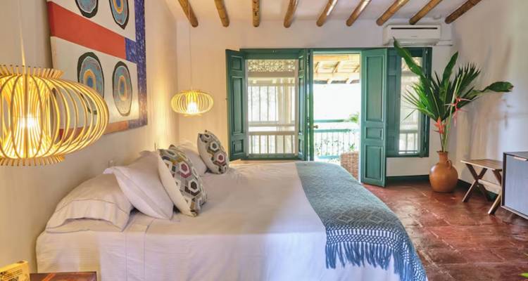 Charmante chambre rustique avec un grand lit soigneusement fait, des poutres de plafond en bois, des suspensions tressées et des volets verts s'ouvrant sur un balcon.