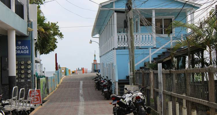 Ruelle étroite en briques menant à un quai, bordée de bâtiments en bois bleu et de motos garées sous des câbles aériens.