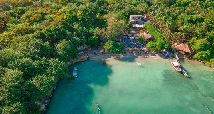 Une plage tropicale isolée avec une eau turquoise, des huttes au toit de chaume et une jungle dense apparaît vue d'en haut.