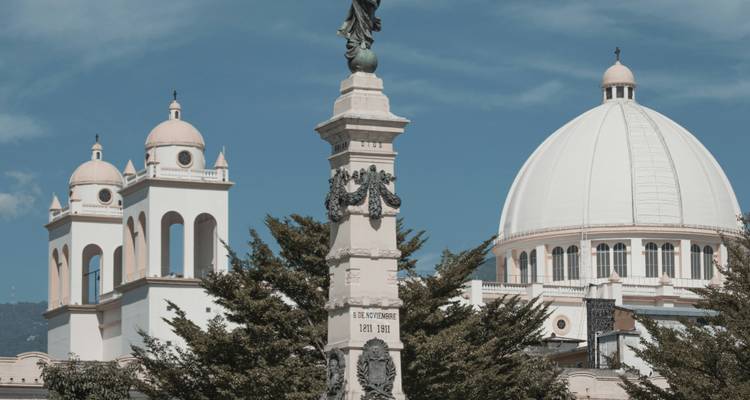 Neoclassical domes and a statue rising above treetops under a clear blue sky in San Salvador.