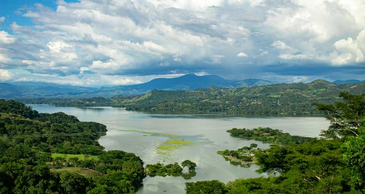 Expansive lake surrounded by lush green hills and distant mountains beneath dramatic clouds.