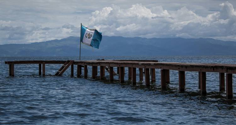 Muelle de madera con bandera guatemalteca ondeando sobre aguas tranquilas del lago y montañas de fondo.