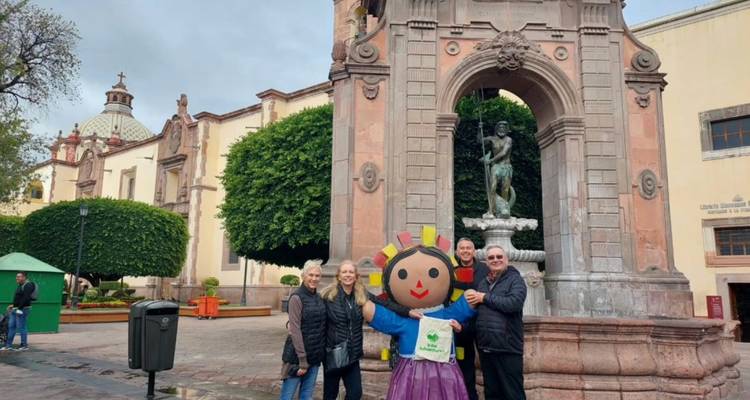 Travelers pose with a large traditional doll in a historic plaza featuring a stone arch and fountain.