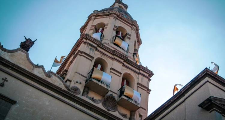 Upward angle of an ornate baroque bell tower adorned with colorful flags against a clear sky.